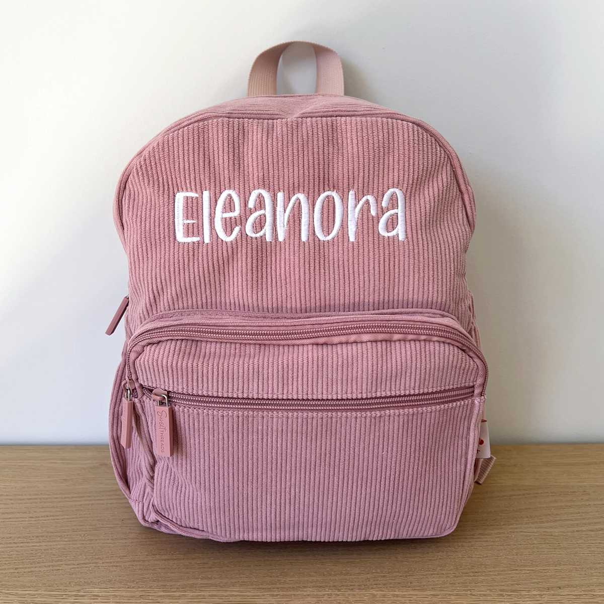 Pink corduroy backpack with 'Eleanora' embroidered on it, placed on a wooden surface with a white background.