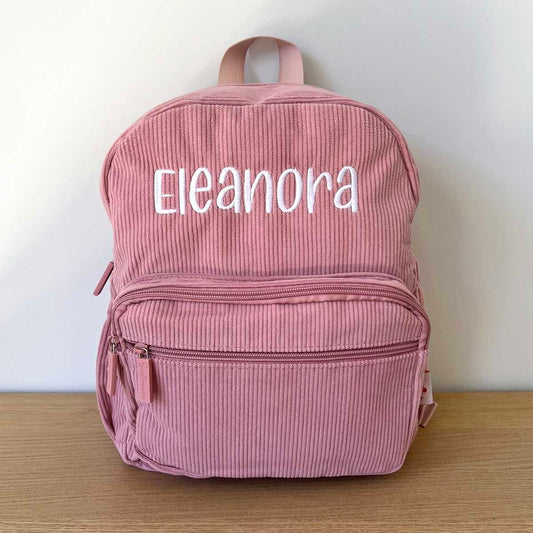 Pink corduroy backpack with 'Eleanora' embroidered on it, placed on a wooden surface with a white background.
