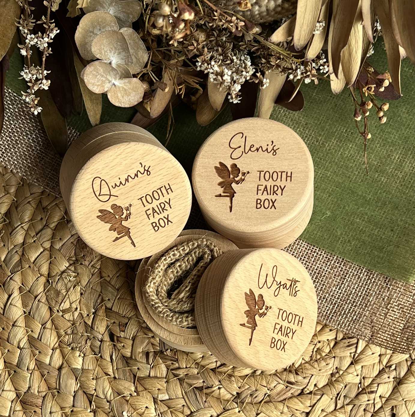 Three wooden tooth fairy boxes with engraved names on a textured surface with flowers and leaves.