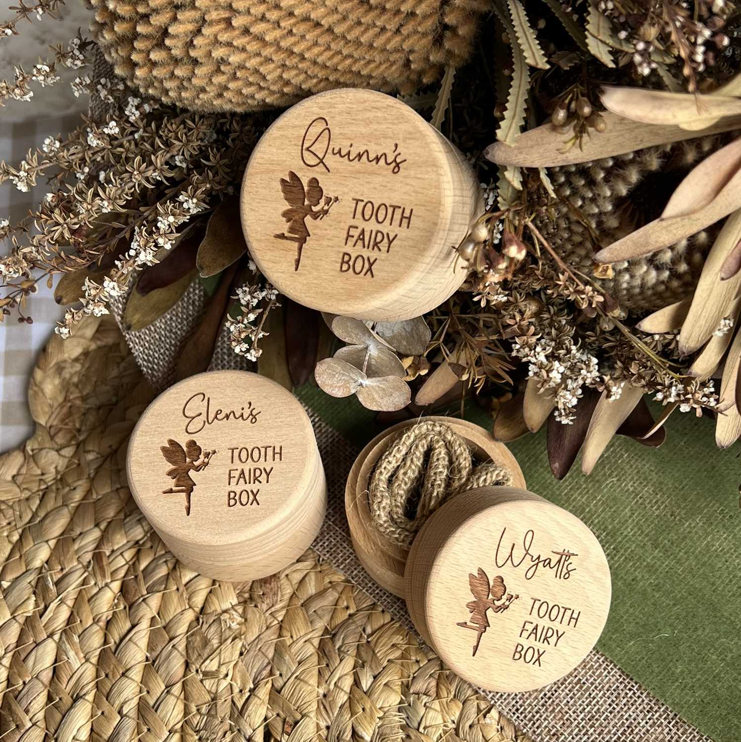 Three wooden tooth fairy boxes with engraved names surrounded by dried flowers and leaves.